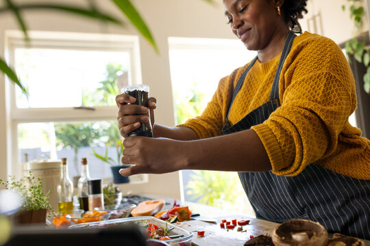 Happy plus size african american woman in apron seasoning vegetables in kitchen