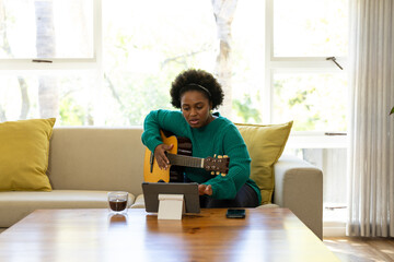 Happy plus size african american woman playing guitar and using tablet at home