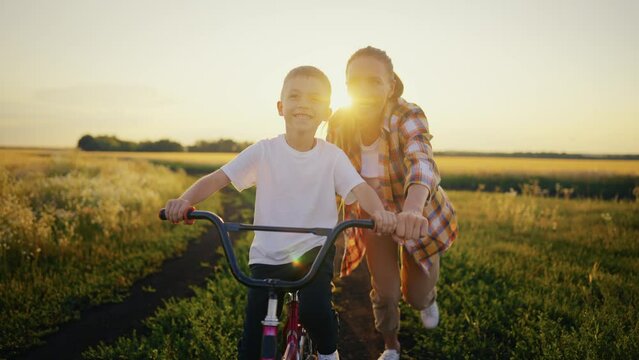 Happy family weekend, smiling mom teaching son riding a bicycle on farm on summer vacation. Mother holding bike with sitting on it joyful child boy. Woman running near. Outdoors activity concept.