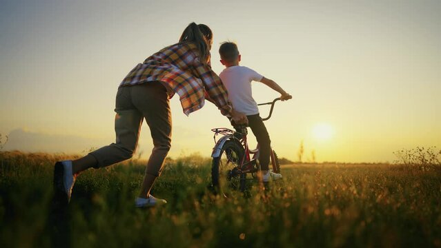 Mom teach son riding bicycle in village on summer vacation between fields on nature at sunset. Mother holding bike with sitting on it child boy. Woman runs after bike. Family walk, weekend outdoors.