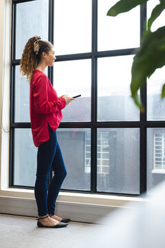 Happy biracial woman holding a smartphone looking out of the window at office