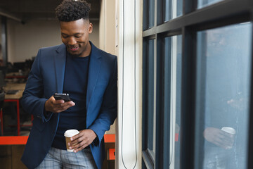 Happy african american man holding a coffee cup using a smartphone at office