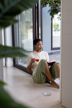 Happy african american woman sitting on floor, drinking tea and reading book