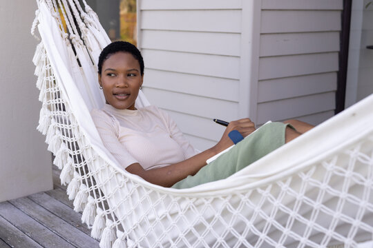 Happy african american woman writing in notebook sitting in hammock on terrace - Powered by Adobe