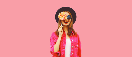 Portrait of happy smiling young woman holding lollipop wearing pink jacket, black round hat on pink background