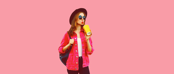 Portrait of stylish young woman drinking fresh juice wearing jacket, black round hat, backpack on pink background