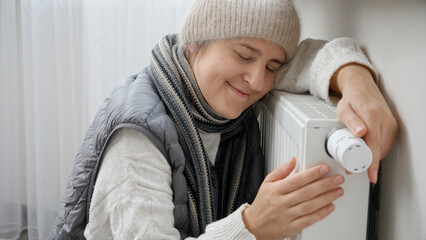 Young woman feeling very cold at home sitting by the heating radiator. Concept of energy crisis,...