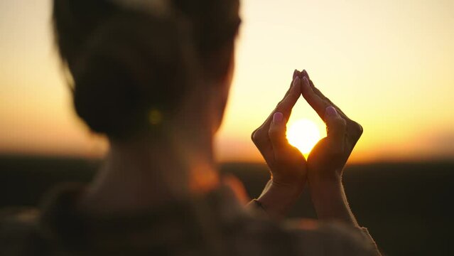 Woman folding hands around sun at sunset in romb shape, symbol of peace and meditation, rear view. Relax, healthy lifestyle, balance, harmony concept. Female in unity with nature, enjoying evening.