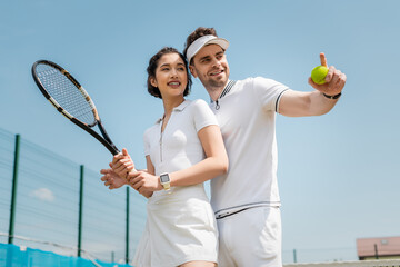 happy man pointing away near girlfriend on tennis court, holding rackets , sport and romance