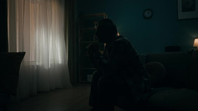 A devastated lonely young man sits in a room in the dark, going through a crisis. The man thinks, does not find a solution, hides his head in his palms. Transience of time, hopelessness. Slow motion.