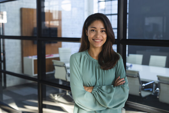 Portrait of happy biracial casual businesswoman with long hair and arms crossed in office