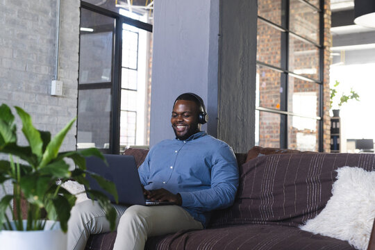 Happy african american casual businessman sitting on couch and using laptop with headphones