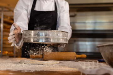 Asian female baker wearing apron in bakery kitchen and pouring flour on dough