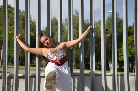 Latina and Hispanic girl, young and pretty, wearing a T-shirt made with a handkerchief and white pants, holding on to a fence with a defiant look. Concept of youth, rebelliousness, defiance, cockiness