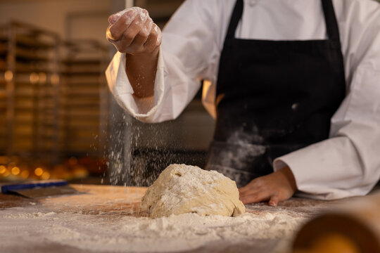 Asian female baker wearing apron in bakery kitchen and pouring flour on dough