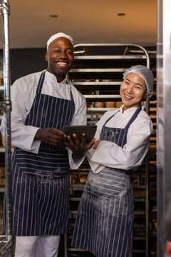 Portrait of happy diverse bakers wearing aprons and using tablet in bakery with arms crossed