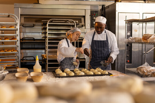 Focused diverse bakers wearing aprons in bakery kitchen and sprinkling seeds on rolls