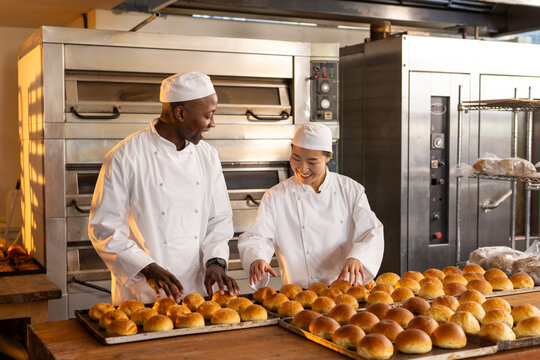 Happy diverse bakers wearing aprons in bakery kitchen and counting fresh rolls