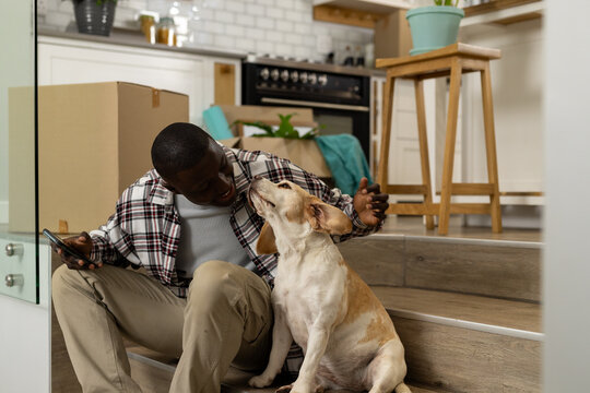 Happy african american man moving house, using smartphone, with his pet dog at home,