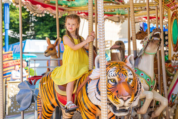 Naklejka na ściany i meble Adorable little blonde girl in summer yellow dress at amusement park having a ride on the merry-go-round. Child girl has fun outdoor on sunny summer day. Entertainment concept