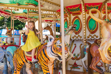 Adorable little blonde girl in summer yellow dress at amusement park having a ride on the merry-go-round. Child girl has fun outdoor on sunny summer day. Entertainment concept
