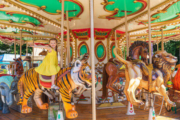Adorable little blonde girl in summer yellow dress at amusement park having a ride on the merry-go-round. Child girl has fun outdoor on sunny summer day. Entertainment concept