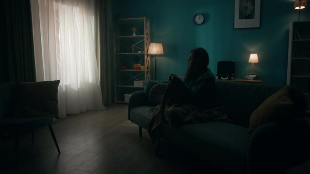 Devastated woman sitting on a sofa in a dark room. A woman desperately thinks about personal problems, is going through a crisis. Change of day and night, transience of time, hopelessness. Slow motion