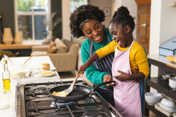 Happy african american mother and daughter baking pancakes in kitchen at home