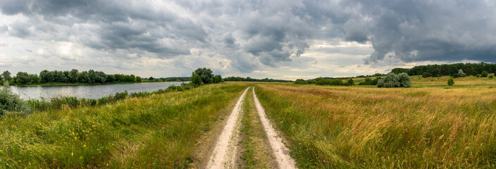 Landscape panorama of a picturesque river valley with a road in a meadow and trees on a cloudy summer day