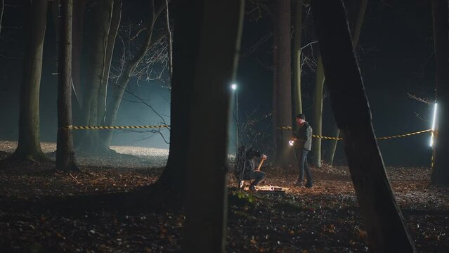Detective and criminologist working on crime scene restricted by police tape in night woods. Glowing flashlight, digging the ground in search of clues. Investigating hard case in spooky setting