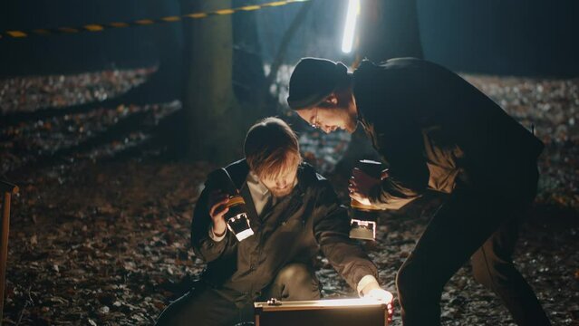 Detective and criminologist working on crime scene in night autumn forest. Close view of investigators looking into open case, laying on the ground. Police work, forensic expert job