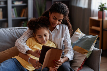 Portrait of loving mother and daughter using digital tablet together relaxing on sofa at home