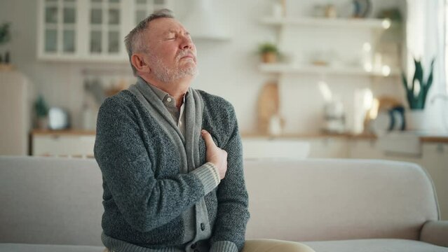 Senior man suffers from heartache sitting on couch at home. Unwell old male with chest pain myocardial infarction has chronic cardiology disease. Cardiac arrhythmia, heart attack, illness concept.