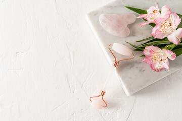 A home quartz roller massager and gua sha scraper lie on a white marble podium stone with flowers. white background. A copy space. natural care
