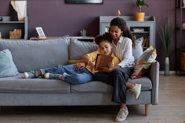 Obraz premium Full length portrait of loving mother and daughter using digital tablet together while relaxing on sofa at home, copy space