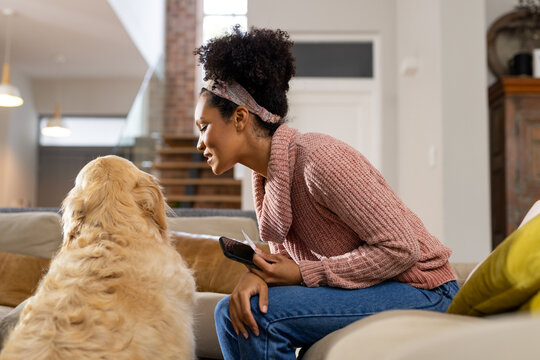 Happy biracial woman petting dog and using smartphone at home