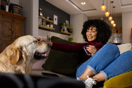 Happy biracial woman petting dog and drinking tea at home