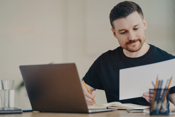 Bearded expert edits reports on laptop, focused at home office, casual in black t-shirt.
