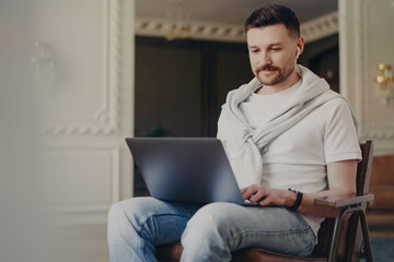 Happy young freelancer, casual clothes, earphones on, using laptop, working from modern home office, listening to music, focused on computer screen.