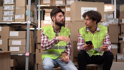 Two warehouse workers in uniform having lunch at workplace watching social networks using smartphone online - Powered by Adobe