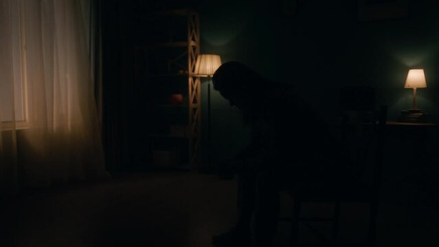 A devastated woman sits with her head bowed on a chair in the middle of the room in pitch darkness. Night follows day, a woman raises her head and looks at the light from the window. Slow motion.