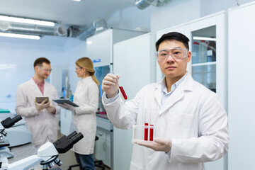 Portrait of a young Asian male doctor, lab technician standing in a lab coat in the clinic and holding tubes with blood, researching for tests, dna. Colleagues are talking in the background.