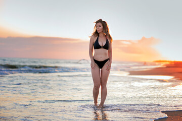 Beautiful brunette woman in a bikini swimsuit on the sandy shore at sunset in warm sunshine
