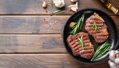 fried beef steaks with rosemary bouquet, garlic and salt on a skillet on a wooden table, horizontal view from above, flat lay, free space