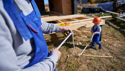 Father with toddler son building wooden frame house. Male builder and kid playing with tape measure...