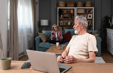 Senior couple argue at home while older retired military man trying to have video call on his laptop computer with army friend and his wife talking to him from behind. Relationship difficulties