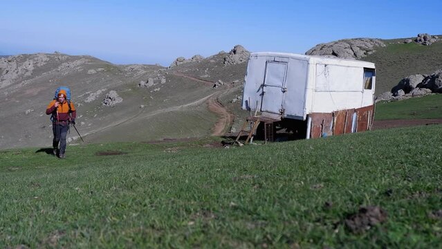Male backpacker walks past trailer bunkhouse toward camera in meadow
