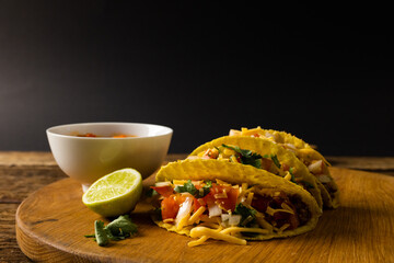 Close-up of tacos with lemon slice, cilantro and bowl on serving board against black background