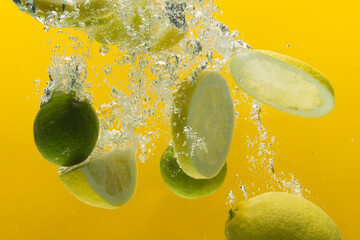 Close up of lemon and lime slices falling into water with copy space on yellow background