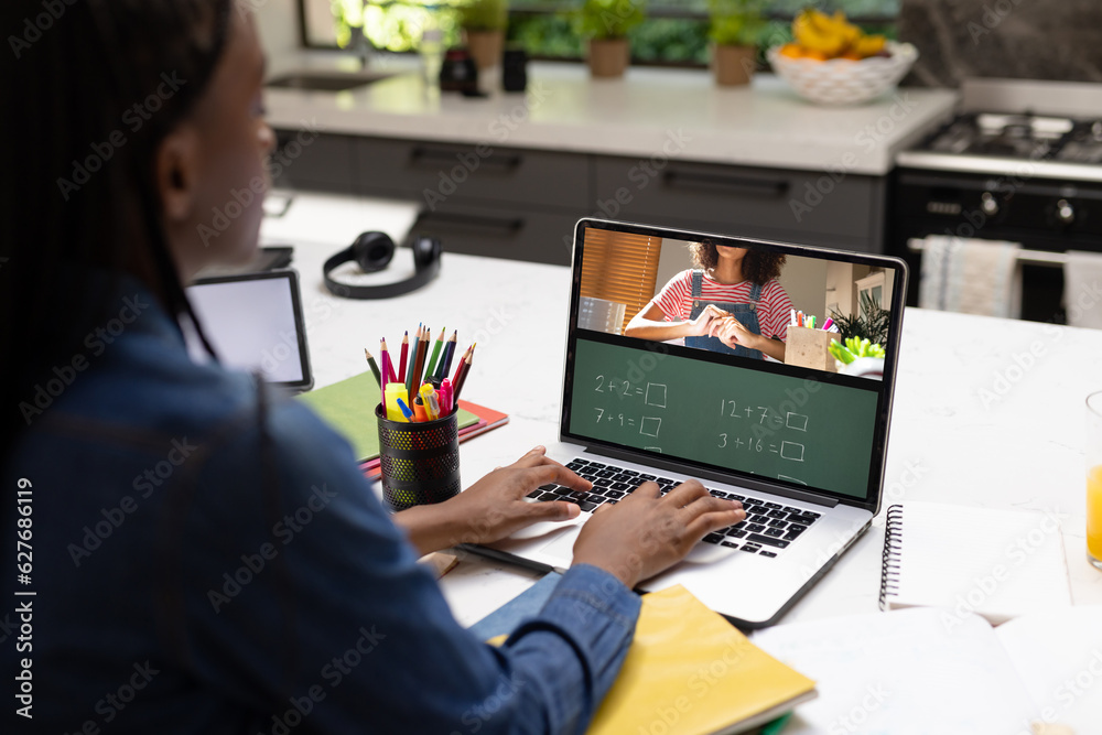 Canvas Prints Rear view of a african american schoolgirl having a video call with female teacher on laptop